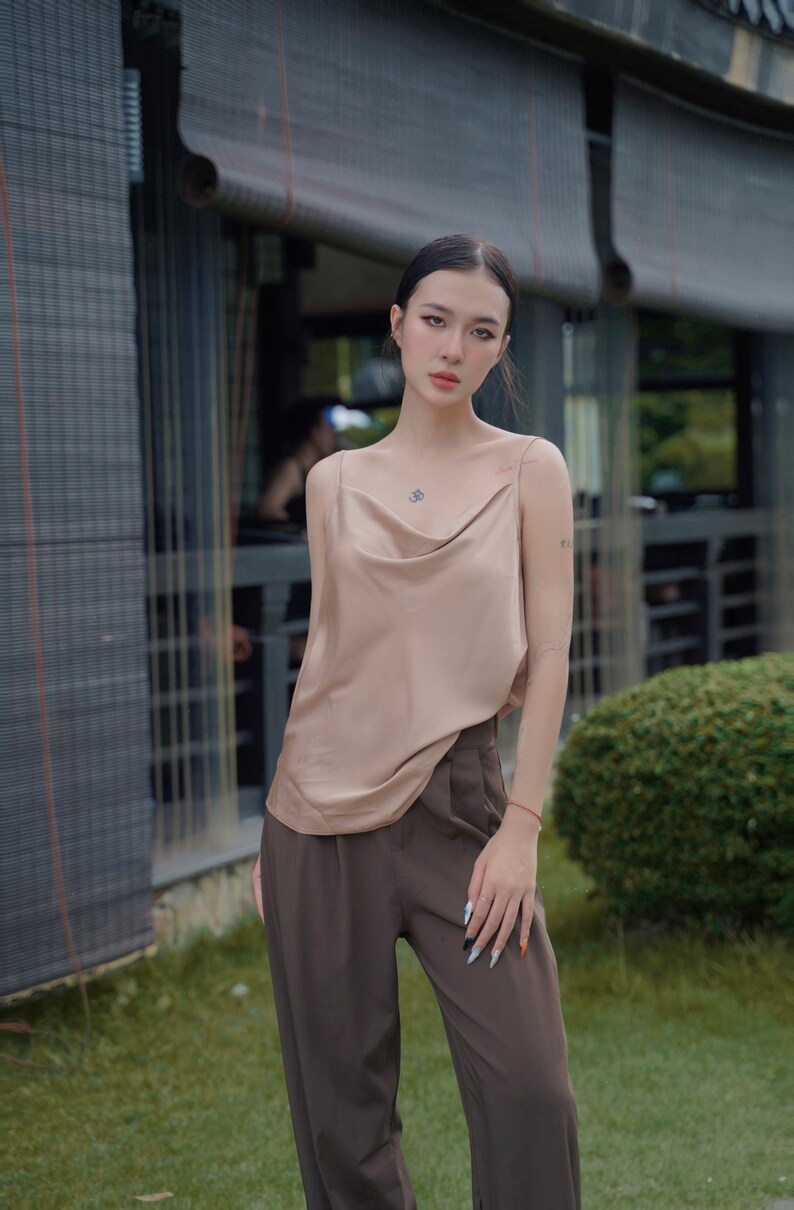 May include: A beige satin camisole featuring a draped cowl neckline and thin spaghetti straps, paired with high-waisted dark brown trousers. The camisole has a smooth, reflective finish, while the trousers feature a tailored fit with visible pleats.