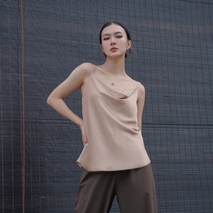 May include: A beige sleeveless camisole top featuring a draped cowl neckline and thin spaghetti straps, paired with dark brown trousers. The top has a smooth, satin-like finish and a relaxed fit, while the trousers are loose-fitting and solid-coloured.