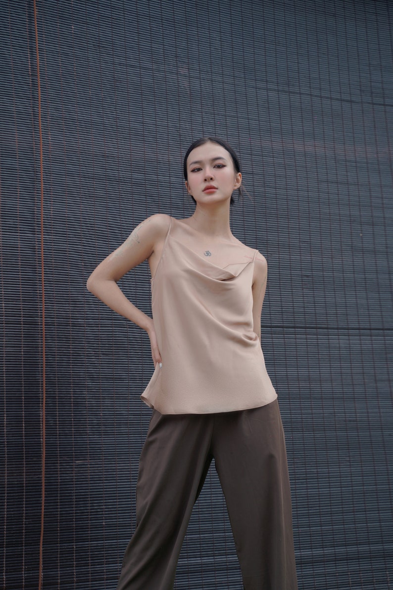 May include: A beige sleeveless camisole top featuring a draped cowl neckline and thin spaghetti straps, paired with dark brown trousers. The top has a smooth, satin-like finish and a relaxed fit, while the trousers are loose-fitting and solid-coloured.