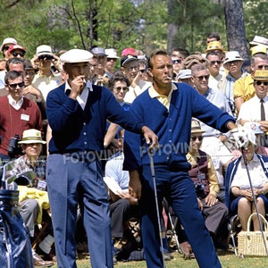 May include: Two men in blue cardigans and slacks stand on a green golf course, surrounded by spectators. One man wears a white cap and the other a yellow collared shirt. Both are smoking cigarettes. The scene is set outdoors on a sunny day.