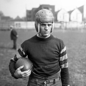 May include: A black and white photo of a football player wearing a leather helmet and a striped jersey. He is holding a football in his left hand and is looking at the camera.
