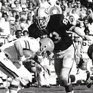 May include: A black and white photo of two football players in action. The player on the left is wearing a white jersey with a brown stripe and a helmet with the word "Bengals" on it. The player on the right is wearing a black jersey with a white number 83 on it and a helmet with the word "Raiders" on it. The players are on a grassy field.