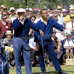 May include: Two men in blue cardigans and slacks stand on a green golf course, holding golf clubs. One man wears a white cap. Spectators in hats and sunglasses watch from behind. The scene is set outdoors on a sunny day.