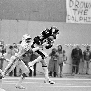 May include: A black and white photo of a football game. A player in a black and white uniform is running with the ball, while two players in white uniforms try to tackle him. The sign in the background reads "Drown the Dolphins".