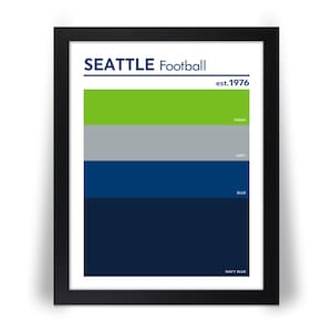 May include: Framed print featuring the text "SEATTLE Football" above a color block design. The design includes green, gray, blue, and navy blue horizontal stripes, with the text "est.1976". The colors are labeled with their names.
