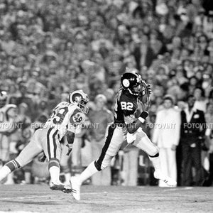 May include: A black and white photo of two football players in action. The player on the left is wearing a white and green uniform with the number 49 on it. The player on the right is wearing a black and white uniform with the number 82 on it. The player on the right is running with the ball and the player on the left is trying to tackle him.