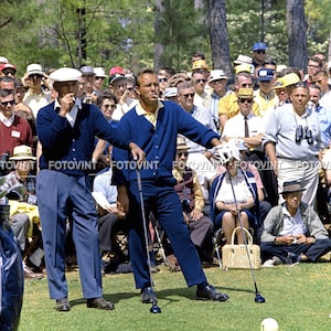 May include: Two men in blue and white clothing stand on a golf course, holding golf clubs. They are surrounded by a crowd of spectators wearing hats and sunglasses.