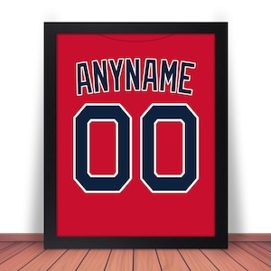 May include: Framed print of a red jersey with the text "ANYNAME" above the number "00" in navy blue with a white outline. The jersey is displayed in a black frame, set against a white wall and a wooden floor.