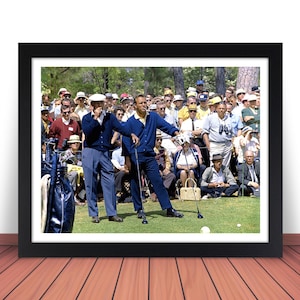 May include: A black and white photograph of two men on a golf course. The man in the foreground is wearing a blue shirt and black pants. He is holding a golf club and is about to swing. The man in the background is wearing a white shirt and black pants. He is watching the man in the foreground.