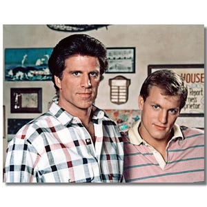 May include: A vintage photograph featuring two men. One man wears a white button-down shirt with a red, black, and blue plaid pattern. The other man wears a pink and white striped polo shirt. The background includes framed artwork and a sign.