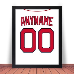 May include: Framed print of a white jersey with the name "ANYNAME" in red, and the number "00" in red with a navy blue outline. The jersey design is set against a white background, framed in black, and displayed on a wooden surface.