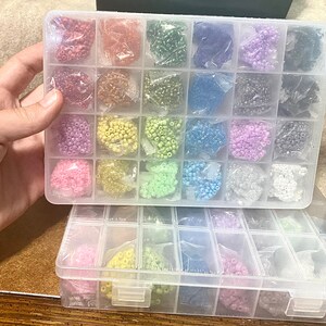 May include: A clear plastic storage box with multiple compartments filled with colorful seed beads. The box is divided into two sections, each with rows of compartments. The beads are arranged in a rainbow pattern.