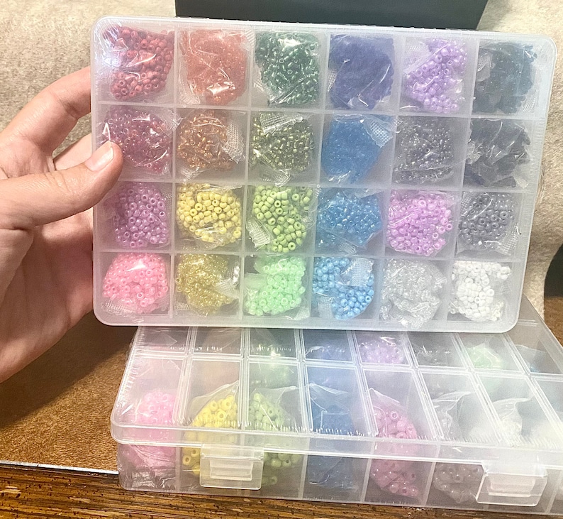 May include: A clear plastic storage box with multiple compartments filled with colorful seed beads. The box is divided into two sections, each with rows of compartments. The beads are arranged in a rainbow pattern.