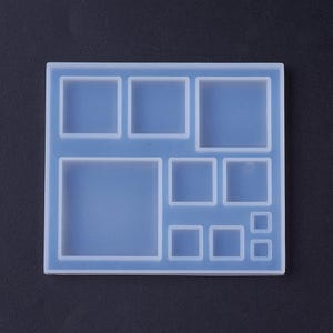 May include: A clear, square silicone mold with various square-shaped cavities. The mold is light blue and has a grid-like pattern of different sized squares. The background is a solid dark gray.