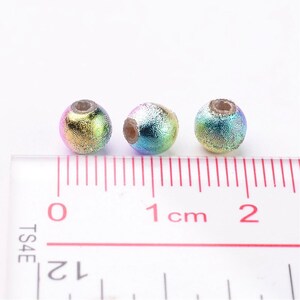May include: Three iridescent round beads with a textured surface, measuring approximately 1 cm in diameter, are lined up on a ruler.