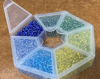 Pode incluir: Um organizador de contas de plástico transparente preenchido com várias cores de pequenas contas de vidro. As cores incluem tons de azul, verde e amarelo. O organizador está aberto, revelando as contas no interior.