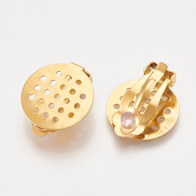 K&ouml;nnte beinhalten: Ein Paar goldfarbene Ohrclips mit rundem, perforiertem Design. Ein Ohrclip hat einen kleinen rosa Stein in der Mitte.