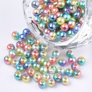 May include: A bowl filled with iridescent round beads in various shades of blue, green, pink, and purple. The beads are scattered on a white surface.