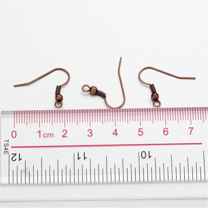 May include: Three copper-colored earring hooks with a small ball at the end of each hook. The hooks are measured against a ruler with markings in centimeters.