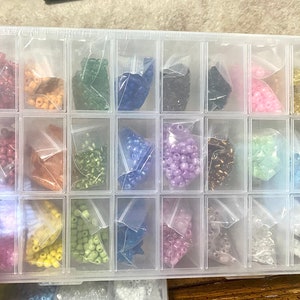 May include: A clear plastic storage box with 18 compartments filled with small bags of colorful beads. The beads are in various colors, including red, orange, yellow, green, blue, purple, pink, and white.