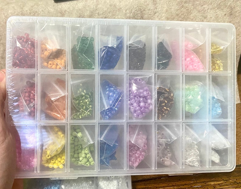 May include: A clear plastic storage box with 18 compartments filled with small bags of colorful beads. The beads are in various colors, including red, orange, yellow, green, blue, purple, pink, and white.