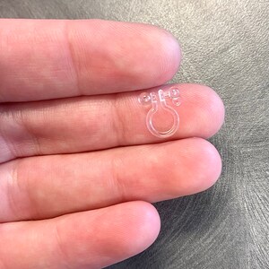 May include: Clear plastic ring with two small bumps on each side. The ring is designed to hold a curtain or other fabric in place.