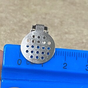 K&ouml;nnte beinhalten: Eine silberfarbene Metall-Filterplatte mit einem Durchmesser von etwa 2,5 cm. Die Filterplatte hat eine runde Form und ist mit einem Gittermuster aus kleinen L&ouml;chern versehen.