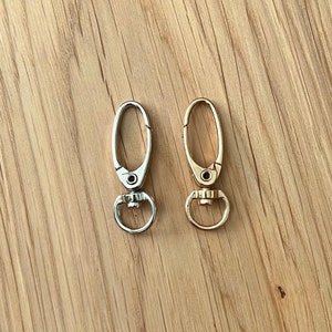 May include: Two metal swivel clasps, one silver and one gold, are displayed against a light brown wood grain background. Each clasp has an oval-shaped hook and a circular ring at the base, designed for attaching to various items.