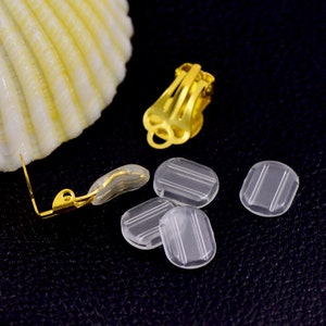 May include: Close-up of gold-colored clip-on earring components and clear, rectangular earring pads. The pads have vertical lines. A gold earring post with a clear pad is also visible. The background is dark.