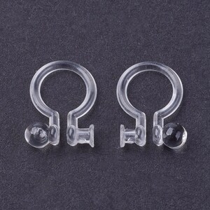 May include: A pair of clear plastic earring backs. The backs have a small, round, clear plastic ball on one end and a small, clear plastic loop on the other end.