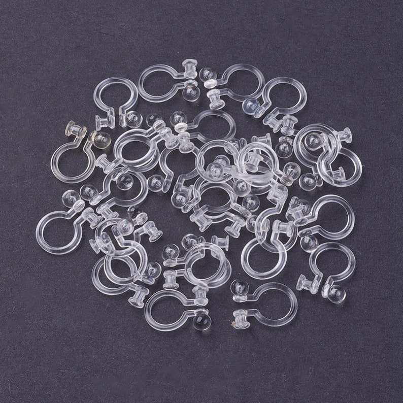 May include: A close-up of a pile of clear plastic earring backs. The backs have a small ball on one end and a hook on the other end.