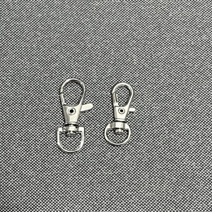 May include: Two silver swivel clasps or hooks. The clasps are closed and are on a gray textured surface.