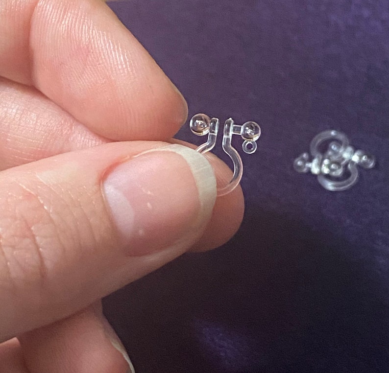 May include: A pair of clear plastic earring backs with small, round, clear beads. The earring backs are designed to be worn on the back of an earring to keep it in place.