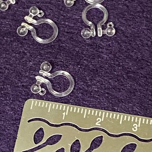 May include: Close-up of clear plastic earring backs with a U-shaped design and small balls on each side. The earring backs are measured against a ruler.