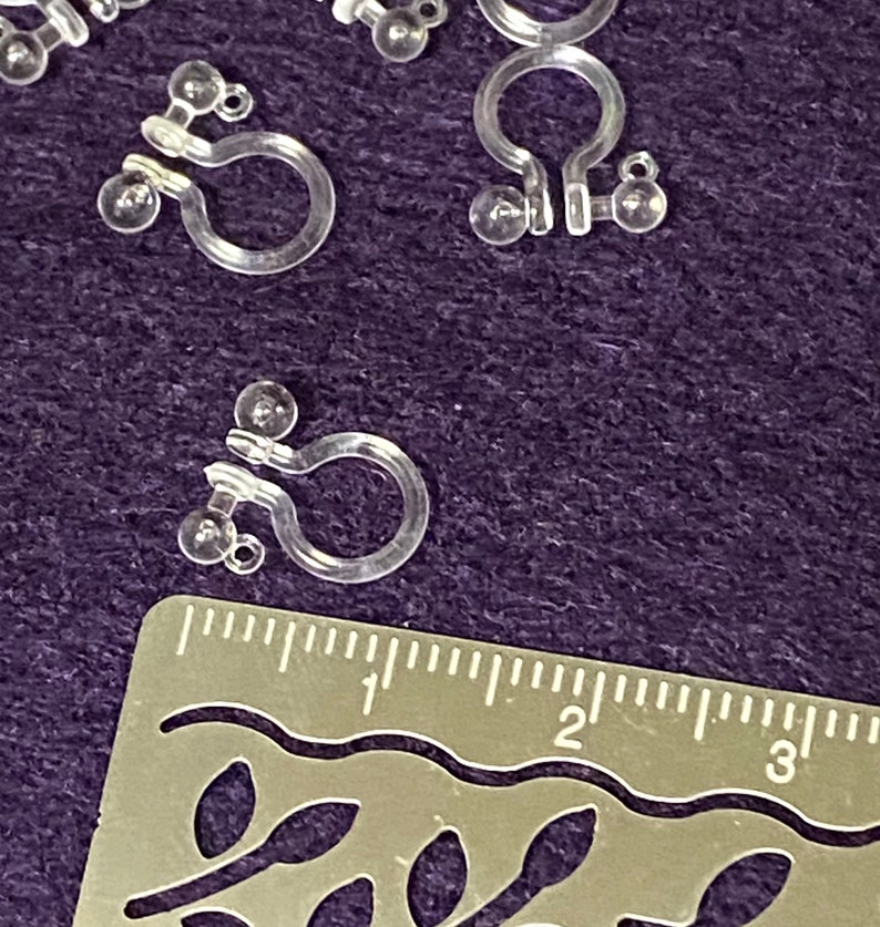 May include: Close-up of clear plastic earring backs with a U-shaped design and small balls on each side. The earring backs are measured against a ruler.