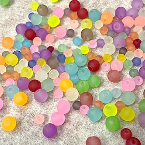 May include: A collection of colorful, translucent beads in various shades of pink, purple, blue, green, yellow, orange, and red. The beads are scattered on a white surface.
