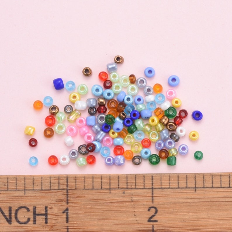 May include: A selection of colourful seed beads in various shades of blue, green, yellow, orange, red, brown, and white. The beads are scattered on a pink surface with a wooden ruler in the foreground.