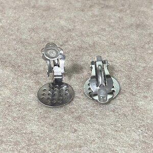 K&ouml;nnte beinhalten: Zwei silberfarbene Ohrring-Clip-Verschl&uuml;sse mit einem runden, perforierten Design. Die Verschl&uuml;sse sind so konzipiert, dass sie einen Cabochon oder andere Verzierungen halten.