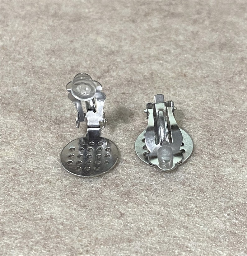 K&ouml;nnte beinhalten: Zwei silberfarbene Ohrring-Clip-Verschl&uuml;sse mit einem runden, perforierten Design. Die Verschl&uuml;sse sind so konzipiert, dass sie einen Cabochon oder andere Verzierungen halten.