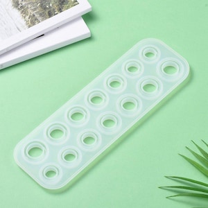 Può includere: Uno stampo in silicone traslucido, rettangolare, con molteplici cavità circolari di varie dimensioni. Lo stampo è progettato per l'artigianato, probabilmente per resina o materiali simili. Lo sfondo è verde chiaro.