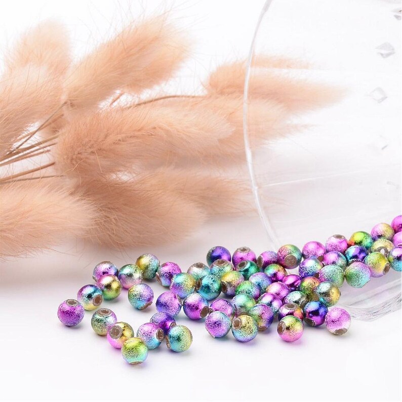May include: A collection of iridescent glass beads in shades of purple, green, and blue. The beads are scattered on a white surface.