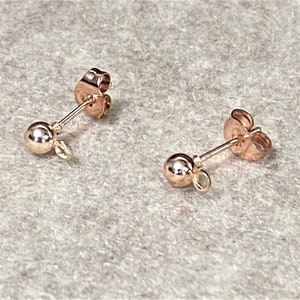 May include: Two small, gold-colored ball stud earrings with post backs. Each earring features a small, round ball and a loop for attaching charms or other decorative elements. The earrings are displayed on a light gray surface.