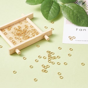 May include: A close-up of a wooden tray filled with gold jump rings. The jump rings are scattered on a light green surface. The text "Fan" and "It's never too late to" are visible on a white card.