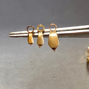May include: Three gold-colored metal pendant bails of different sizes, held by a pair of tweezers. The bails are shaped like a teardrop with a round loop at the top.