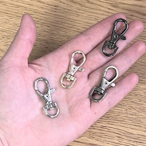 May include: Four metal swivel clasps in silver, gold, and gunmetal gray are displayed in an open hand. The clasps have a spring-loaded gate and a circular loop for attaching to a strap or other item. The background is a light wood surface.