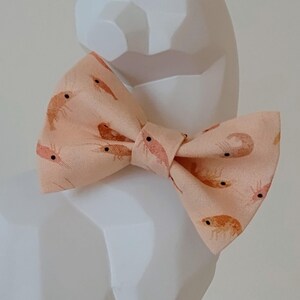 Peut inclure: Un nœud papillon pêche avec un motif répété de crevettes de dessin animé. Le nœud papillon est noué dans un style classique et est fait de tissu. Les crevettes sont orange avec des yeux noirs.