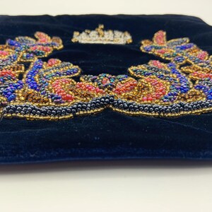 May include: A black velvet pouch with a beaded design of a crown surrounded by leaves. The beads are in shades of red, blue, gold, and iridescent.
