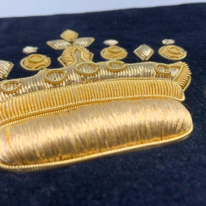 May include: A gold embroidered crown on a dark blue fabric background. The crown has a rounded top and is decorated with gold thread.