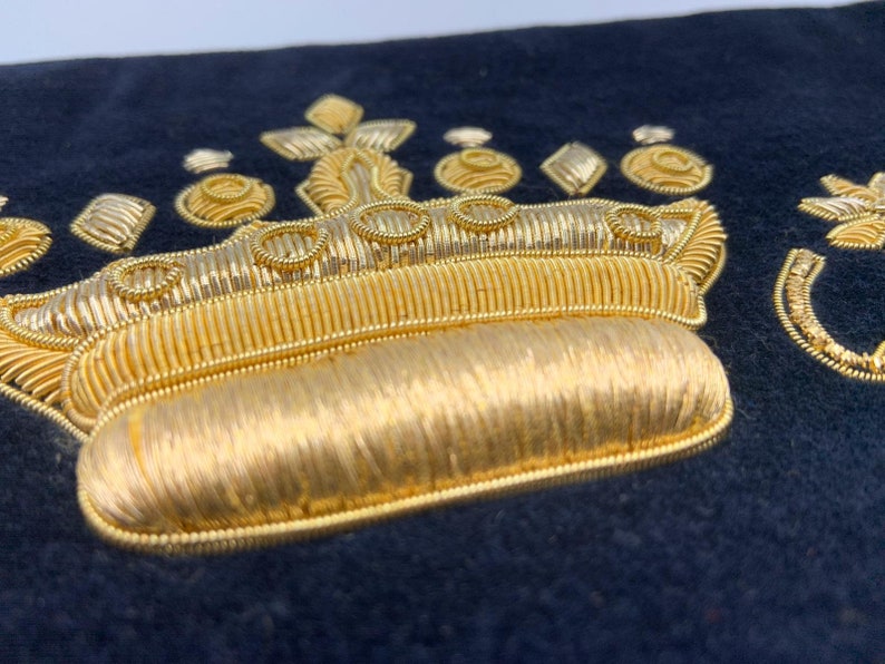 May include: A gold embroidered crown on a dark blue fabric background. The crown has a rounded top and is decorated with gold thread.