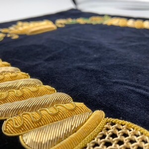 May include: A black velvet cushion with gold embroidery. The embroidery features a crown, a chain, and other decorative elements.
